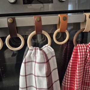 Pode incluir: Um conjunto de suportes para toalhas de cozinha com anéis de madeira e tiras de couro. Os suportes estão presos a uma barra de metal, com toalhas xadrez vermelhas e brancas penduradas. As tiras de couro são em várias tonalidades de castanho.