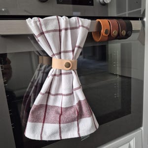Può includere: Un canovaccio bianco con motivo a quadri rosso scuro, fissato da un cinturino in pelle color cuoio chiaro con un bottone a pressione in ottone. Tre cinturini in pelle aggiuntivi in cuoio, marrone e marrone scuro pendono dalla maniglia del forno.