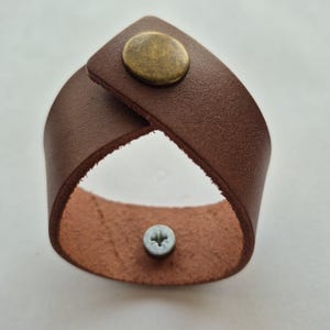 May include: A brown leather bracelet with a brass-colored button closure and a silver screw. The bracelet is made of a single strip of leather that crosses over itself.