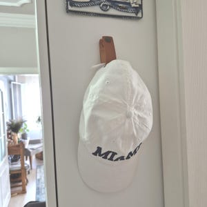 May include: A white baseball cap with the word MIAMI embroidered in dark blue lettering on the front. The cap is suspended from a brown leather strap attached to a vertical surface, featuring a button detail at the top of the crown.