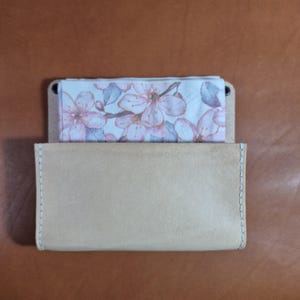 Könnte beinhalten: Ein hellbrauner Lederkartenhalter mit einem floral bedruckten Einsatz. Der Kartenhalter ist rechteckig mit einer genähten Kante. Der Einsatz zeigt rosa und weiße Kirschblüten auf hellblauem Hintergrund.