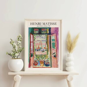 K&ouml;nnte beinhalten: Ein gerahmter Druck eines Gem&auml;ldes von Henri Matisse mit dem Titel "Offenes Fenster, Collioure, 1905". Das Gem&auml;lde zeigt eine farbenfrohe Ansicht eines Fensters mit einer offenen T&uuml;r, die auf einen Hafen mit Booten blickt. Das Gem&auml;lde befindet sich in einem wei&szlig;en Rahmen.