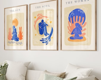 Könnte beinhalten: Set aus drei gerahmten Kunstdrucken mit abstrakten Illustrationen. Jeder Druck hat einen beigen Hintergrund mit blauen und orangefarbenen Figuren. Der Text auf den Drucken lautet "THE SUN", "THE SOUL" und "THE WOMAN". Die Rahmen sind aus hellbraunem Holz.