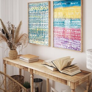 May include: Two framed abstract art prints with colorful geometric patterns. The prints are hung on a white wall above a wooden console table with books and a vase of dried flowers.