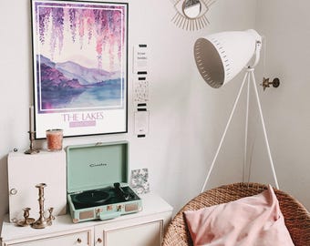 May include: A mint green record player with a black turntable sits on a white dresser with a framed watercolor print of a purple mountain landscape with the text "THE LAKES" and a pink candle. A white lamp with a tripod stand is in the background.