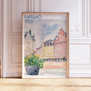 Puede incluir: Impresión en acuarela de Malmö, Suecia, con edificios coloridos y una maceta en primer plano. La impresión incluye el texto "MALMÖ" y "Sweden" en azul. La obra de arte está enmarcada en un marco de madera clara.