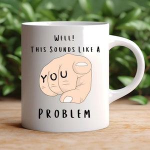 Könnte beinhalten: Weiße Keramik-Kaffeetasse mit einer schwarz-weißen Illustration eines zeigenden Fingers mit dem Wort "YOU" darauf. Der Text "WELL! THIS SOUNDS LIKE A PROBLEM" ist auf die Tasse gedruckt.