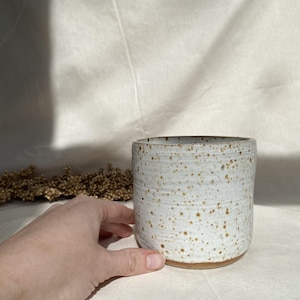 Op de afbeelding: Een gespikkelde witte keramische beker met een bruine basis. De cilindrische beker heeft een getextureerd oppervlak en wordt met een hand in het frame getoond. De beker is waarschijnlijk handgemaakt en geschikt voor dranken.