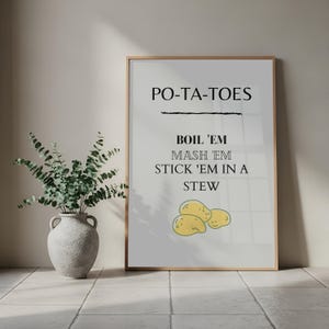 Könnte beinhalten: Ein gerahmter Druck mit dem Text "PO-TA-TOES" über den Worten "BOIL 'EM, MASH 'EM, STICK 'EM IN A STEW". Der Druck enthält eine gelbe Illustration von drei Kartoffeln. Links befindet sich eine dekorative Vase mit grünem Laub.