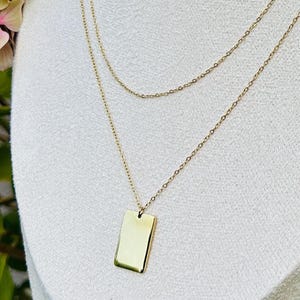 May include: A gold-toned layered necklace featuring two delicate chains of different lengths. The longer chain holds a small, smooth, rectangular pendant with rounded corners. The metal has a polished, reflective finish throughout the entire piece.