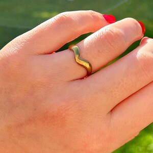 May include: A gold-coloured ring with a wavy design. The ring is on a person's finger, with a green background. The ring's design is a continuous wave, adding a touch of elegance to the jewellery.