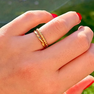 May include: Two gold rings on a finger. One is a plain band, the other has a beaded design. The rings are worn on a hand with red nail polish. The background is blurred green.