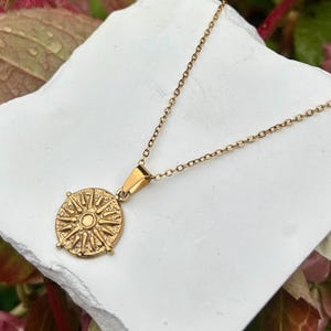 Peut inclure: Un collier doré avec un pendentif circulaire représentant un motif de soleil. Le pendentif est suspendu à une chaîne délicate. Le collier est présenté sur une surface blanche texturée, avec un arrière-plan flou de feuillage vert et rose.