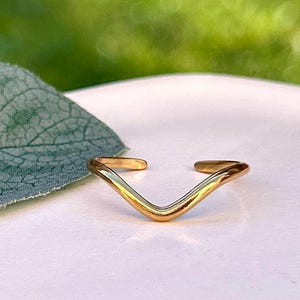 May include: A gold-coloured, open-ended ring in a V-shape. The ring is set against a white surface with a blurred green background and a large green leaf. The ring's design is simple and elegant.