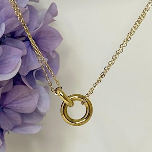 May include: A gold-colored necklace with a circular pendant. The chain is delicate and the pendant is a simple, polished ring. The background features soft purple flowers and a white surface.