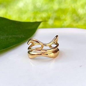 May include: A gold ring with an unusual, open design. The ring has a series of curved, flowing lines that create an abstract shape. The ring is set against a white surface with a blurred green background.
