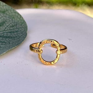 Könnte beinhalten: Ein goldfarbener Ring mit offenem ovalem Design. Der Ring hat eine strukturierte Oberfläche und ist vor einem weißen Hintergrund mit verschwommenem grünem Hintergrund platziert.