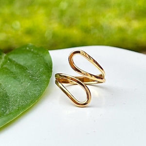 May include: A gold ring with a distinctive open design. The ring has two overlapping teardrop shapes, giving it an elegant and modern appearance. The ring is set against a white surface with a green leaf and blurred green background.