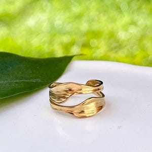 May include: A gold ring with a textured, layered design. The ring has a flowing, organic shape, with two bands that appear to overlap. The background is a soft green, suggesting a natural setting. The ring is likely made of metal.
