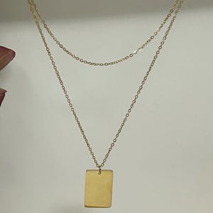 Puede incluir: Un collar dorado en capas con un colgante rectangular. El collar presenta dos delicadas cadenas, con el colgante colgando de la cadena inferior. El fondo es de un color suave y neutro, y se ven unos pétalos de flores moradas a la izquierda.
