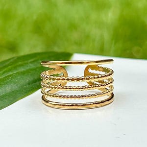 Puede incluir: Un anillo dorado con un diseño multicapa. El anillo presenta varias bandas, incluyendo elementos lisos, con cuentas y texturizados. El anillo se muestra sobre una superficie blanca con un fondo verde, resaltando sus intrincados detalles y brillo metálico.