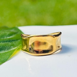 May include: A gold ring with a rectangular face and curved edges. The ring has a polished, reflective surface, and is set against a blurred green and white background. The ring appears to be made of metal.