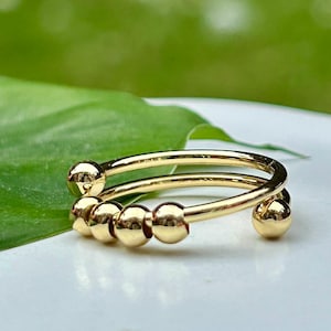 Könnte beinhalten: Ein goldfarbener Ring mit einem einzigartigen Design. Der Ring hat zwei Bänder mit kleinen, runden Perlen, die sich frei bewegen. Der Ring ist vor einem verschwommenen grünen Hintergrund platziert, der sein glänzendes, metallisches Finish hervorhebt. Der Ring ist ein Schmuckstück.