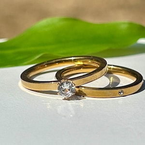 May include: Two gold-toned rings with a smooth, polished finish. One ring features a single, round, clear faceted stone set in a prong setting. The second ring is a simple band with a single, small, flush-set clear stone embedded in the metal.
