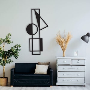 Könnte beinhalten: Eine schwarze Wandskulptur aus Metall mit geometrischen Formen, darunter ein Kreis, Quadrate und Dreiecke, vor einer weißen Wand. Die Skulptur hängt über einem schwarzen Sofa mit einem weißen Kissen, einer grauen Kommode und einer Topfpflanze.