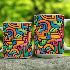 May include: Two white ceramic mugs with colorful abstract designs. The mugs are different sizes and are sitting on a gray stone surface.