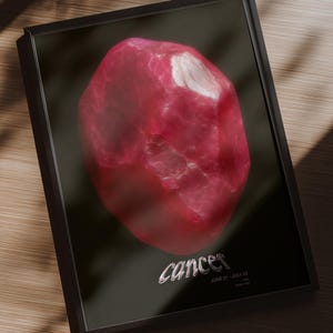 Op de afbeelding: Ingelijste print met een grote, getextureerde, roze edelsteen tegen een zwarte achtergrond. Het woord "Cancer" is in zilveren letters geschreven, met de data 21 juni - 22 juli eronder. De print zit in een zwarte lijst.