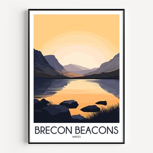 May include: A framed art print featuring a landscape illustration of the Brecon Beacons in Wales. The scene depicts dark mountains flanking a calm lake reflecting a pale yellow sunset. The text BRECON BEACONS WALES is printed at the bottom.