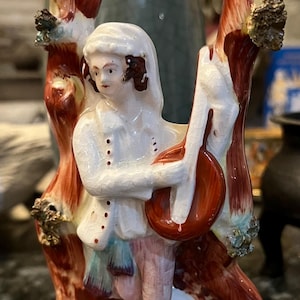 Könnte beinhalten: Eine Porzellanfigur einer Person, die ein Musikinstrument spielt, stehend in einem Baum mit roten und goldenen Akzenten. Die Figur ist weiß mit bemalten Details.