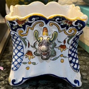 May include: A white ceramic planter with a blue and yellow floral design. The planter has a square shape with rounded edges and a raised, three-dimensional dragon head in the centre.