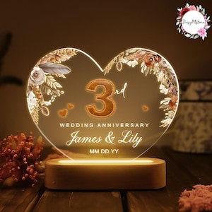 May include: A heart-shaped 3rd wedding anniversary LED light. The clear acrylic design features floral accents and the names "James & Lily." The base is made of wood. The light is illuminated with a warm glow.