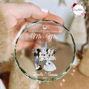 Può includere: Un ornamento natalizio in vetro circolare e sfaccettato con il testo stampato "OUR FIRST CHRISTMAS AS Mr + Mrs" sopra un'illustrazione di Topolino e Minnie in abiti da sposa davanti a un castello, con "Davis + Sarah" e "2025" in basso.