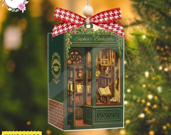 May include: A green and brown bookstore ornament with the text "Sophia's Bookstore". The ornament has a red and white bow and a wooden bead at the top. The ornament is hanging from a string. The words "Name Can Be Customized" are at the bottom.
