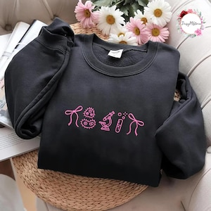 Pode incluir: Camisola preta com gráficos bordados em rosa com tema científico e a palavra "RESIN". O design inclui um microscópio, um tubo de ensaio e outros símbolos científicos. A camisola é exibida numa superfície tecida.