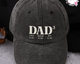 May include: A charcoal gray baseball cap with the word "DAD³" in white block letters. Below the word "DAD³" are the names Lisa, David, and Ace, with their established years. The cap has a curved brim and a fabric strap.