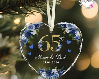 May include: A heart-shaped glass ornament featuring a gold glitter 65th anniversary design, blue floral accents, and small blue hearts. The text reads 65th, Mum & Dad, and 05.08.2026. It hangs from a white ribbon against a blurred evergreen tree background.