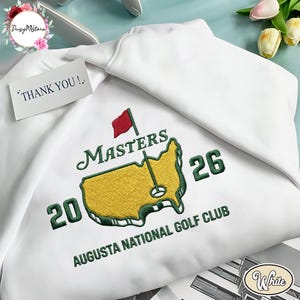 May include: White sweatshirt with embroidered "Masters 26" logo featuring a golf course map and a red flag. The text "Augusta National Golf Club" is below the logo. A small tag reads "THANK YOU!".