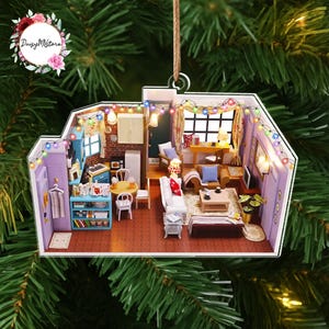 May include: A detailed, miniature diorama ornament depicting a living space with a kitchen, living room, and dining area. The ornament features a string of colorful lights, a brick wall, and various furniture pieces. The ornament is hanging from a brown string.