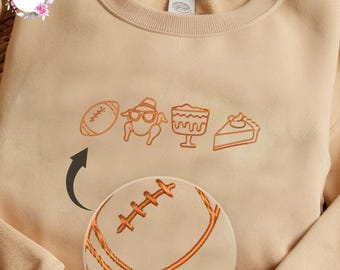 May include: Beige sweatshirt with orange embroidered Thanksgiving-themed graphics. The design includes a football, a turkey wearing glasses and a hat, a bowl of dessert, and a slice of pie. A close-up of the football is also shown.