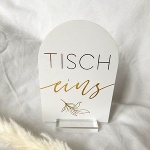 Könnte beinhalten: Weißes Tischnummernschild mit goldener Beschriftung "Tisch eins" und einem goldenen Blattdesign. Das Schild steht auf einem durchsichtigen Acrylständer.