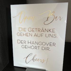 Może przedstawiać: Biały znak akrylowy z pozłacanymi napisami: "Open Bar", "Die Getränke gehen auf uns", "Der Hangover gehört dir" i "Cheers!"