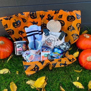 Könnte beinhalten: Ein Halloween-Geschenkkorb in orangefarbenem Papier mit schwarzen Geister- und Kürbismotiven, gefüllt mit einem weißen Becher, weißen Socken mit schwarzen Streifen, einem Beutel mit Geisterillustration, Pringles, Trolli-Süßigkeiten und einem kleinen gehäkelten Kürbis.