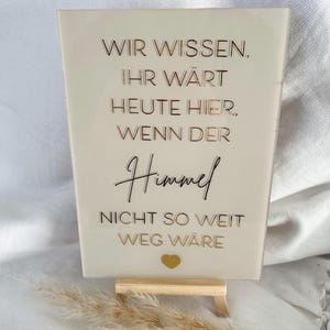 Może przedstawiać: Biały znak akrylowy z pozłacanymi literami, na kt&oacute;rym widnieje napis "Wir wissen, Ihr w&auml;rt heute hier, wenn der Himmel nicht so weit weg w&auml;re". Znak stoi na małym drewnianym stojaku i ma złote serce w prawym dolnym rogu.