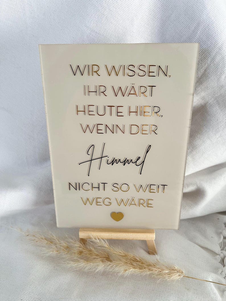 Może przedstawiać: Biały znak akrylowy z pozłacanymi literami, na kt&oacute;rym widnieje napis "Wir wissen, Ihr w&auml;rt heute hier, wenn der Himmel nicht so weit weg w&auml;re". Znak stoi na małym drewnianym stojaku i ma złote serce w prawym dolnym rogu.