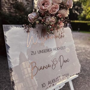 Könnte beinhalten: Ein klares Acrylschild für Hochzeiten mit Aquarellhintergrund und roségoldenen Lettern. Das Schild lautet "Willkommen zu unserer Hochzeit Bianca & Dane 10. August 2024".