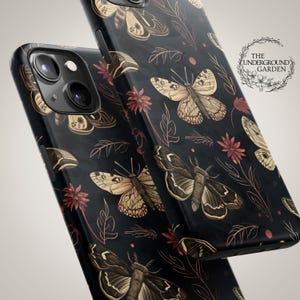 Puede incluir: Carcasa de teléfono negra con un patrón repetido de polillas y flores. La carcasa tiene un fondo oscuro con detalles dorados y rojos. El texto "The Underground Garden" está impreso en la carcasa.